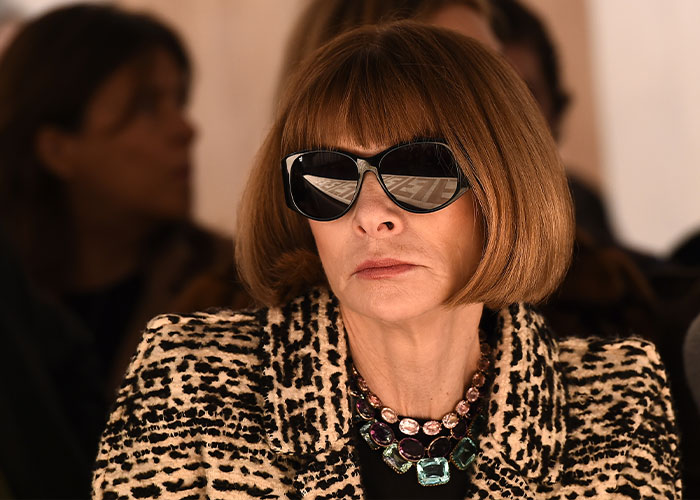 Anna Wintour wearing large sunglasses and a leopard print coat at a fashion event, embodying Vogue icon status.