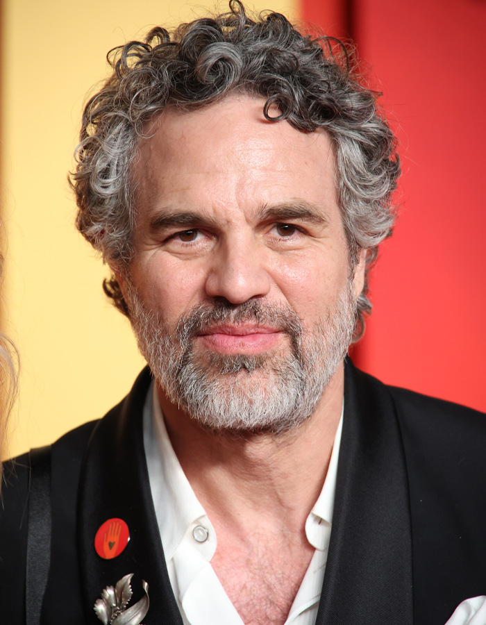 Mark Ruffalo in a close-up portrait with gray hair and beard, wearing a black jacket and white shirt at a red event. Mark Ruffalo in a close-up portrait with gray hair and beard, wearing a black jacket and white shirt at a red event.