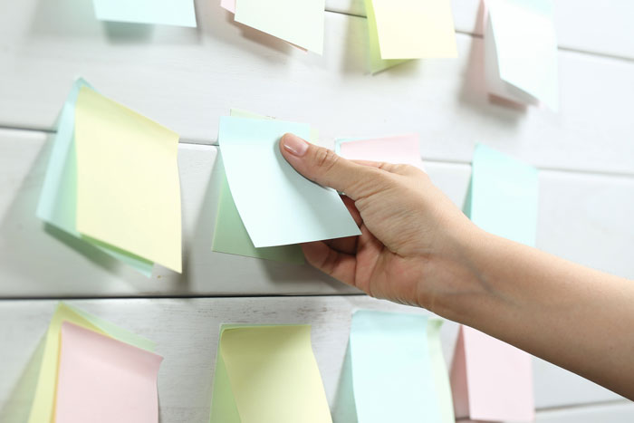 Hand placing a blank pastel sticky note on a wall covered with multiple colorful notes, illustrating petty karma at work.
