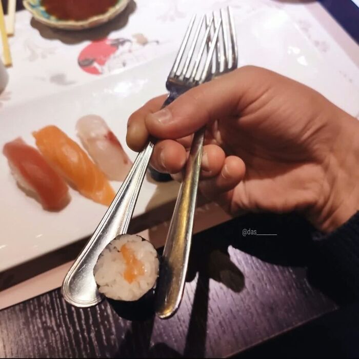 A person awkwardly holding sushi with two forks, a truly weird image. More sushi rests on a plate nearby.
