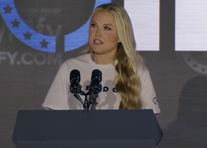 Erika Kirk falando em um pódio durante uma polêmica visita a uma escola secundária em meio à revolta adolescente e à reação pública.