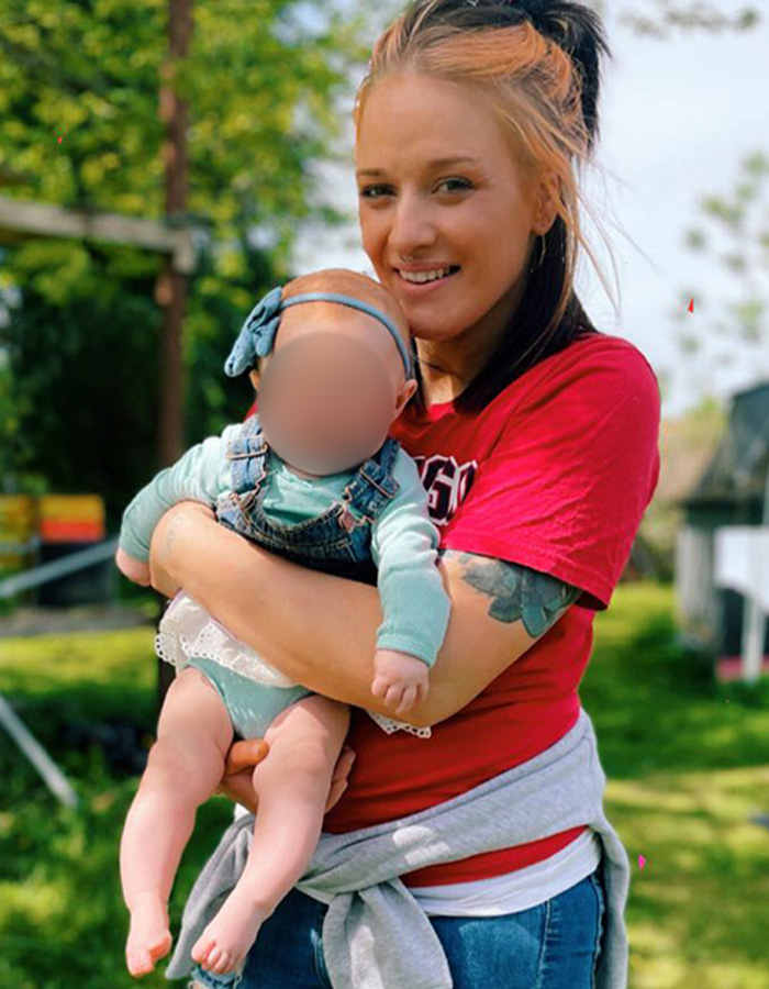 Jovem segurando bebê ao ar livre em uma área verde, relacionado aos restos mortais de uma mãe desaparecida encontrada por caminhantes depois de mais de um ano.