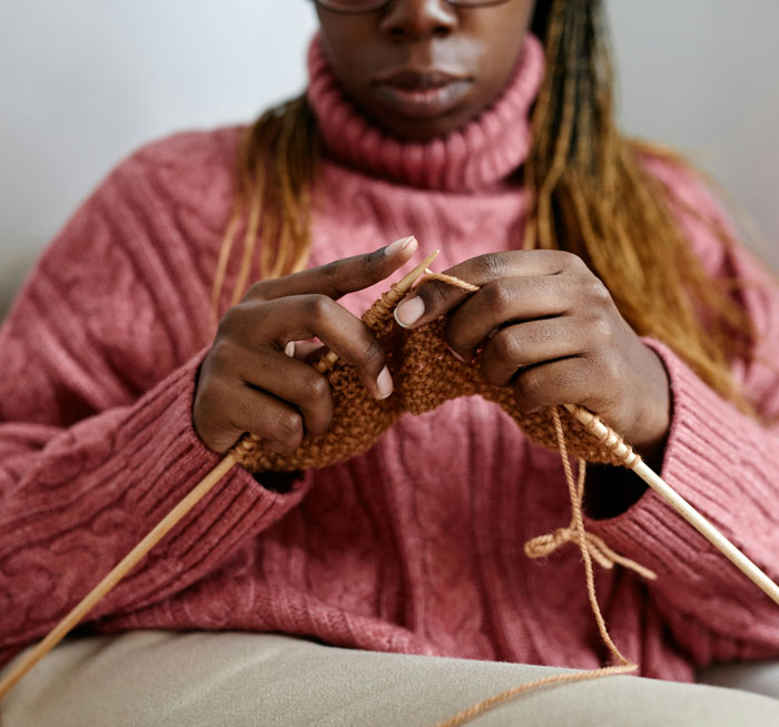 A person knitting with dark skin, wearing a pink sweater and glasses, enjoying a hobby. Favorite things about Black people.