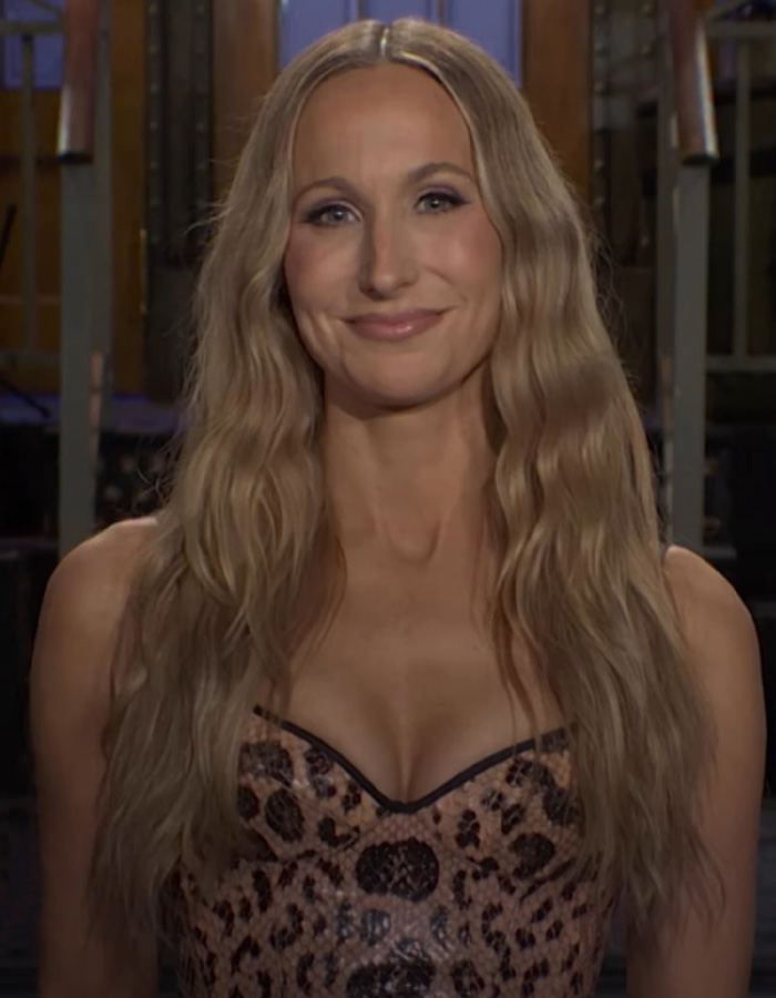 Nikki Glaser com longos cabelos loiros, sorrindo levemente, vestindo um top com estampa de leopardo. Ela se dirige ao rosto sério característico de Victoria Beckham.