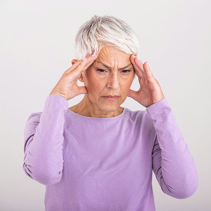 Uma mulher idosa, de cabelos grisalhos, vestindo uma camisa roxa, segura as têmporas, expressando desconforto. Tratamento de Alzheimer para ovos.