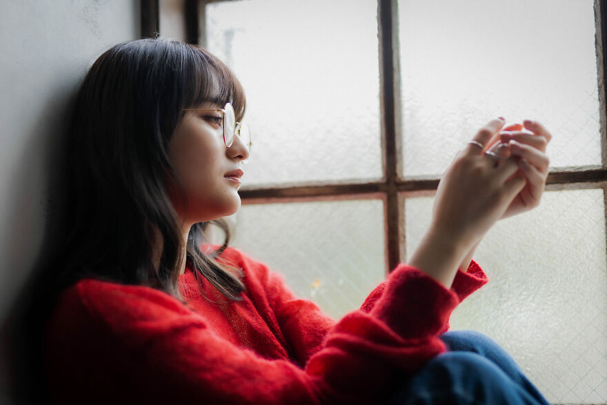 A young woman in a red sweater and glasses looks out a window, reflecting on financial a***e in marriage.