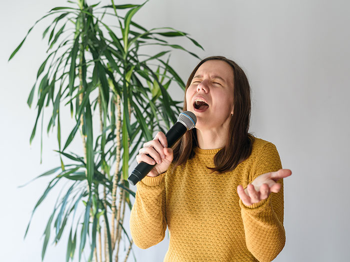 A woman with brown hair, wearing a yellow sweater, sings passionately into a microphone. This may be how she feels after horrible first dates.