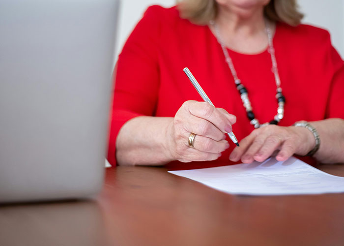 Woman in red shirt writing on paper at desk next to laptop, illustrating signs of the economy not doing well.