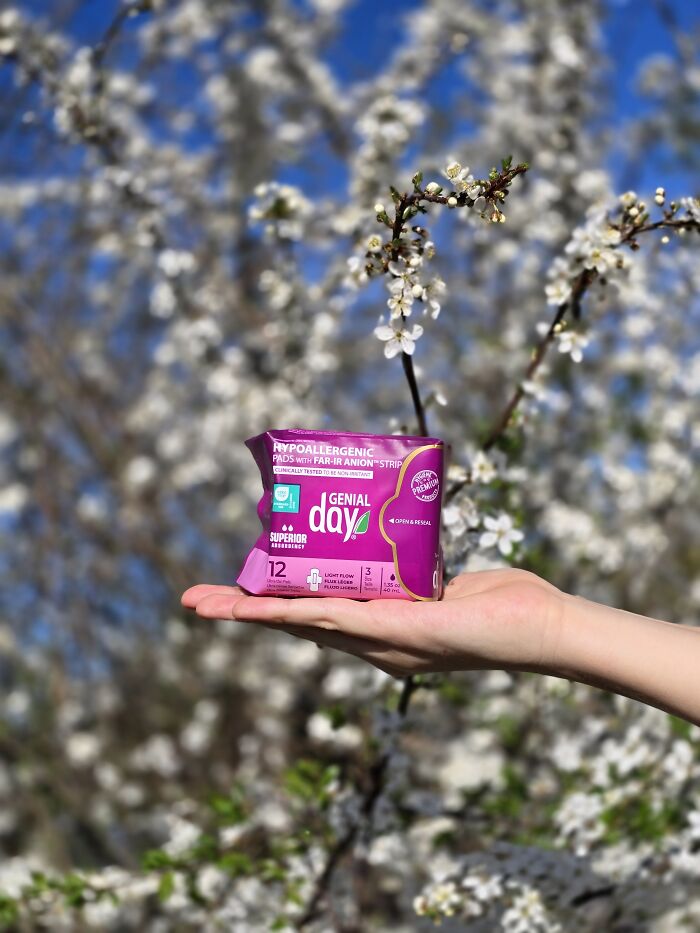 A hand holding a pack of Genial Day pads, with a blurred background of white blossoming trees. Advice for dads raising daughters.