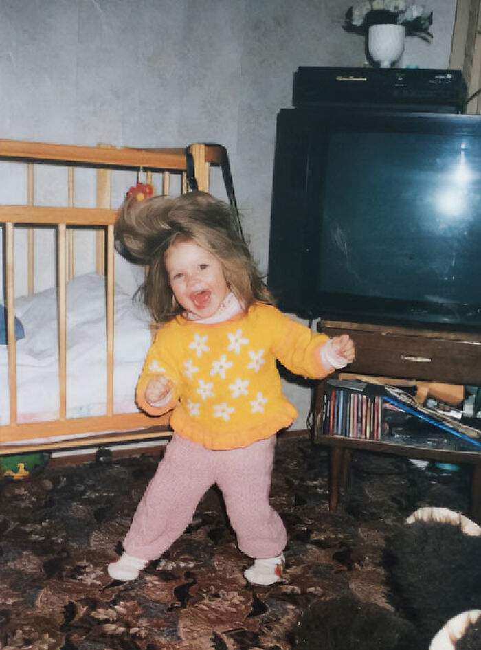 Child dancing wildly in a colorful sweater in a chaotic childhood photo with a crib and old TV in the background.