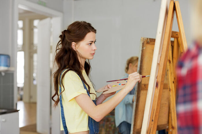 Young woman artist painting intently, her thoughtful expression hinting at deep reflection on friendship betrayals.