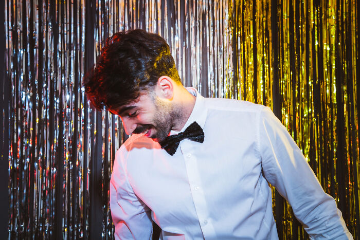 Man in white shirt and black bow tie laughing at a wedding reception with shiny silver and gold backdrop decorations.
