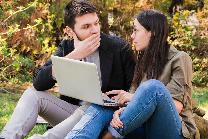 A man covering his mouth in shock as a woman shows him something on a laptop. They are sharing wildest rumors.