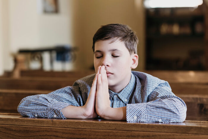 A boy in a plaid shirt with closed eyes, praying at a church pew, contemplating leaving the church.