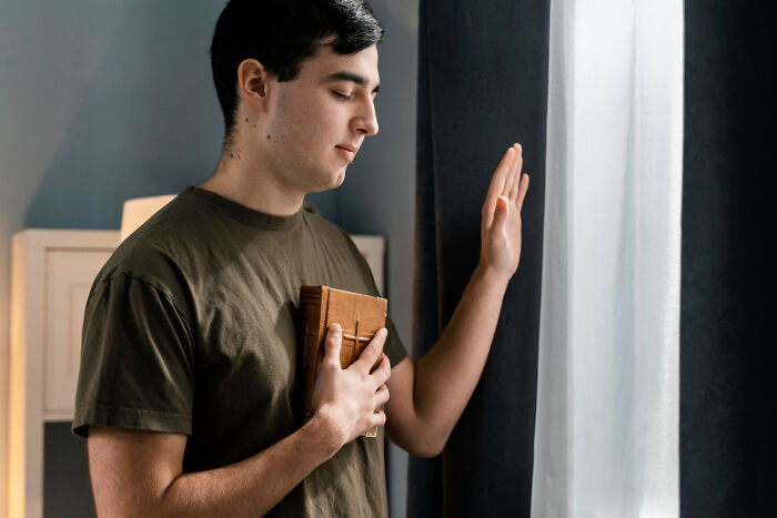 A young man with closed eyes, holding a book to his chest and raising a hand, a former priest contemplating faith.
