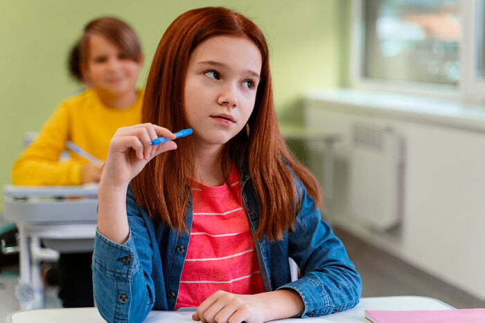 A thoughtful red-haired girl in a denim shirt ponders, holding a blue pen, potentially hearing wild rumors.