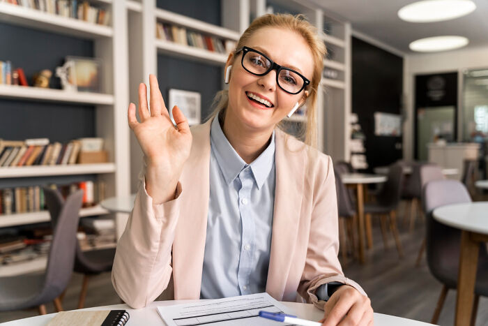 Smiling woman in glasses waving during a virtual meeting, illustrating jobs that AI can’t touch in modern workplaces.