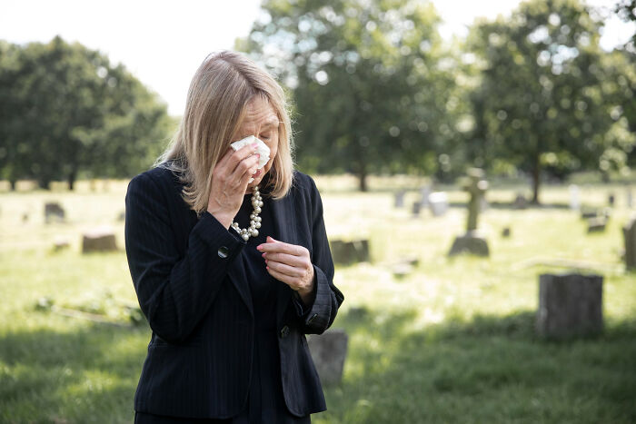 A distraught woman in a black suit with a pearl necklace cries and wipes her eyes with a tissue in a graveyard. Disturbing sounds.