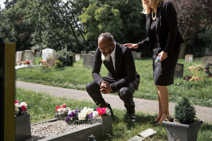 A man in a suit crouches by a grave, being comforted by a woman. They mourn amidst whispers of wild rumours.