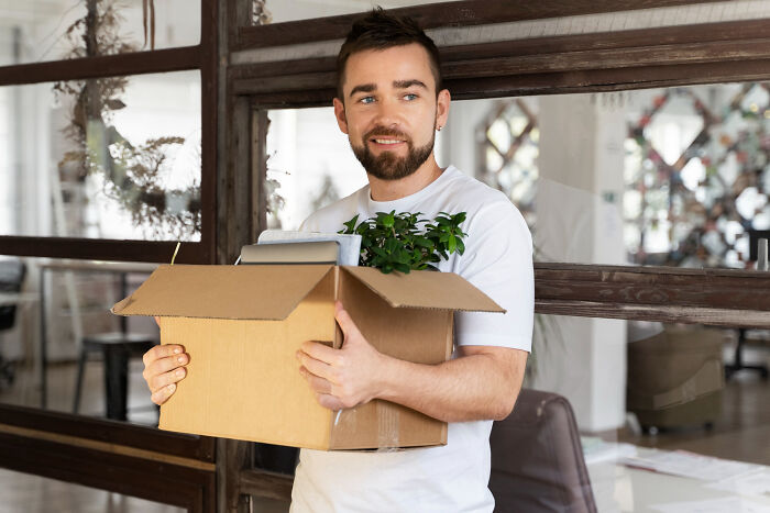 Smiling bearded man holds a moving box with a plant, signaling a fresh start after friendship betrayals.