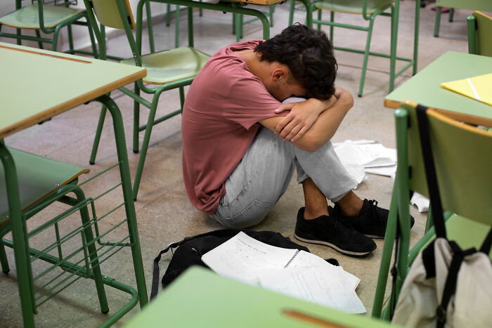 A person sits curled on the floor amidst school desks, head in arms, appearing distressed, dealing with wildest rumours.
