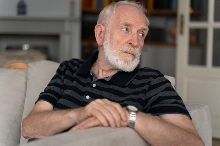 Elderly man with white beard sitting on couch, looking away thoughtfully, depicting kidney donation refusal situation.