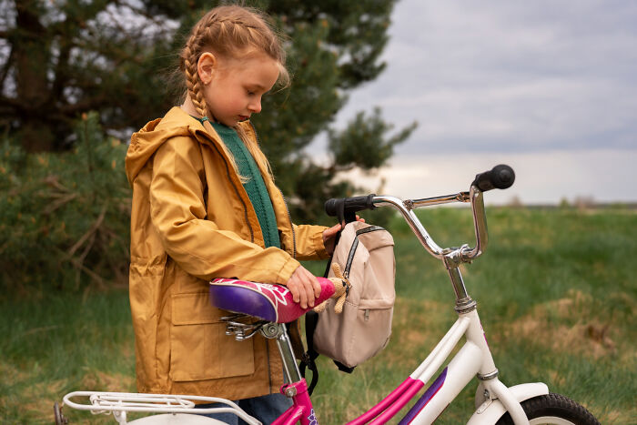 Young girl with braids and a yellow coat holding a backpack on a bike, illustrating kids’ lack of survival instincts.