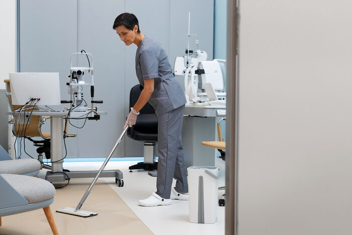 A medical worker in grey scrubs mops the floor of a clean examination room with eye exam equipment.