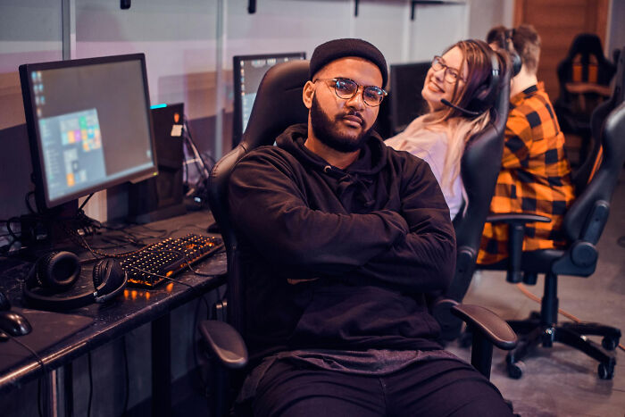 Man wearing glasses and a beanie sitting confidently in a gaming chair with others nearby showing satisfying karma moments.