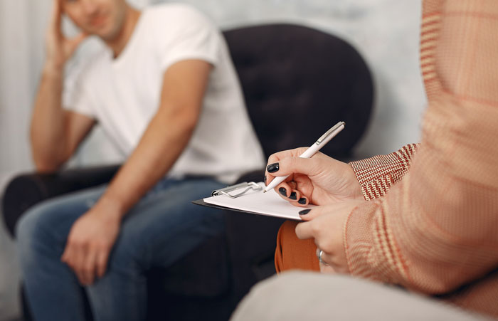 Man in casual clothes looking distressed during therapy session with counselor taking notes on clipboard about family issues