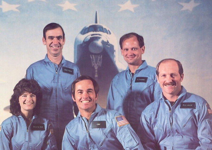 Five astronauts in blue flight suits posing in front of a space shuttle representing events from 1960 to 2010.