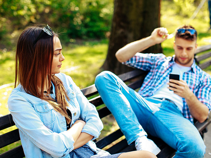 A woman with crossed arms looks annoyed while a man on a bench is engrossed in his phone, symbolizing horrible first dates.