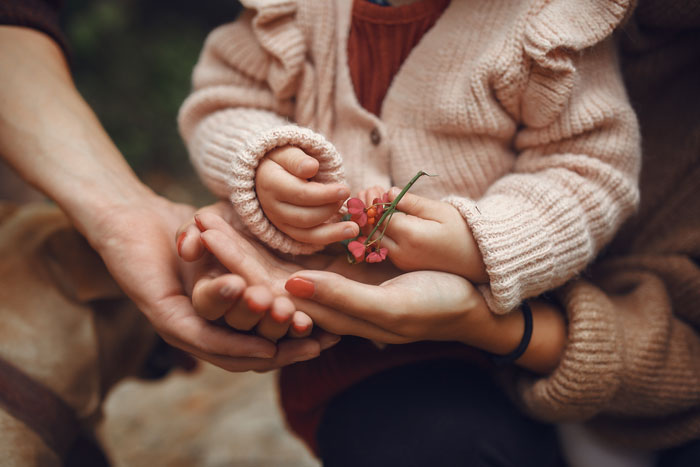 Child holding small flowers with mother and father’s hands surrounding, symbolizing family dynamics with kids.