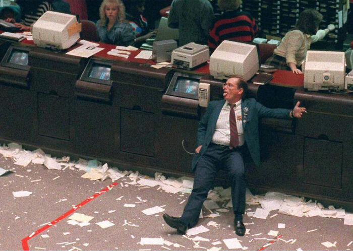 Trader sitting on floor in busy stock exchange surrounded by paper chaos, illustrating key moments from the year you were born.