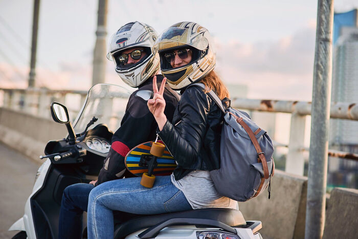 Two helmeted riders on a scooter, the passenger giving a peace sign. A skateboard is attached. Medical Workers Witnessed.