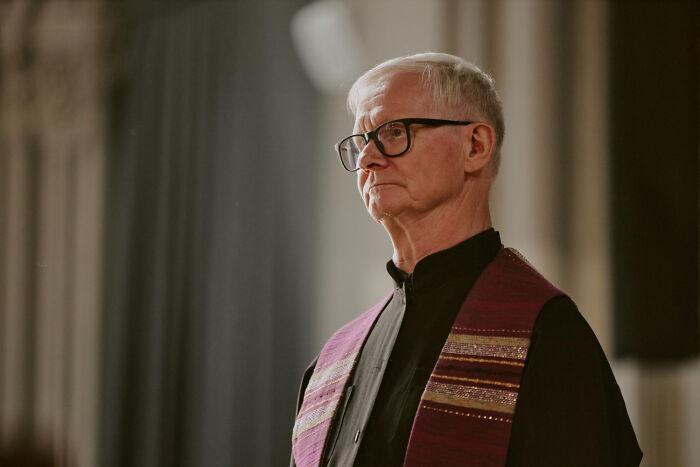 Elderly man wearing glasses and a clerical robe, reflecting on moments of being a good person and betrayal.