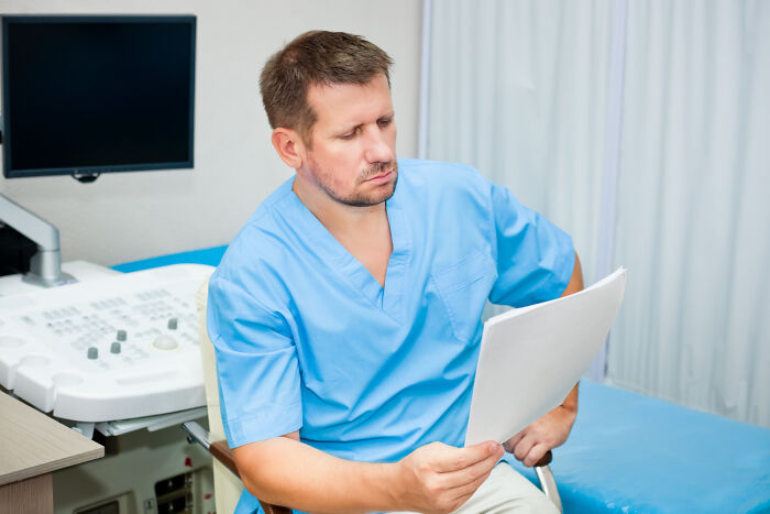 Male gynecologist in blue scrubs reviewing patient documents thoughtfully in a medical office setting.