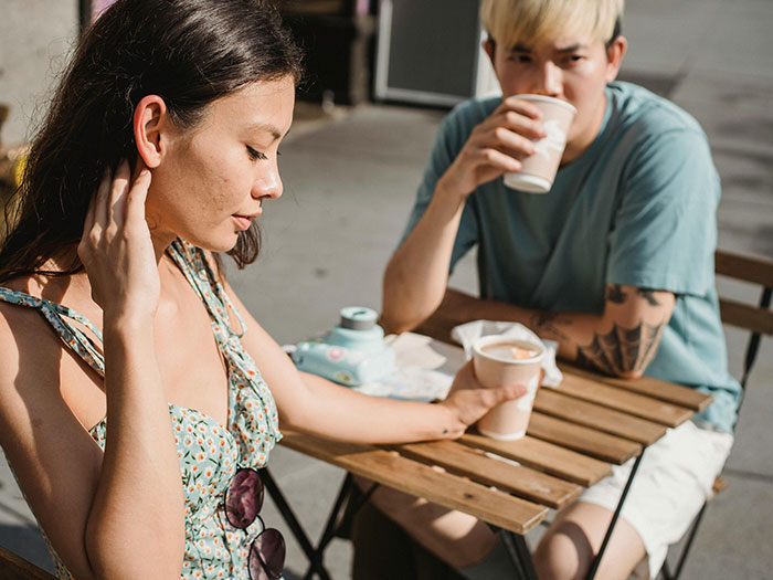 A woman looking down, holding a cup, and a man drinking coffee outdoors. Illustrates horrible first dates.