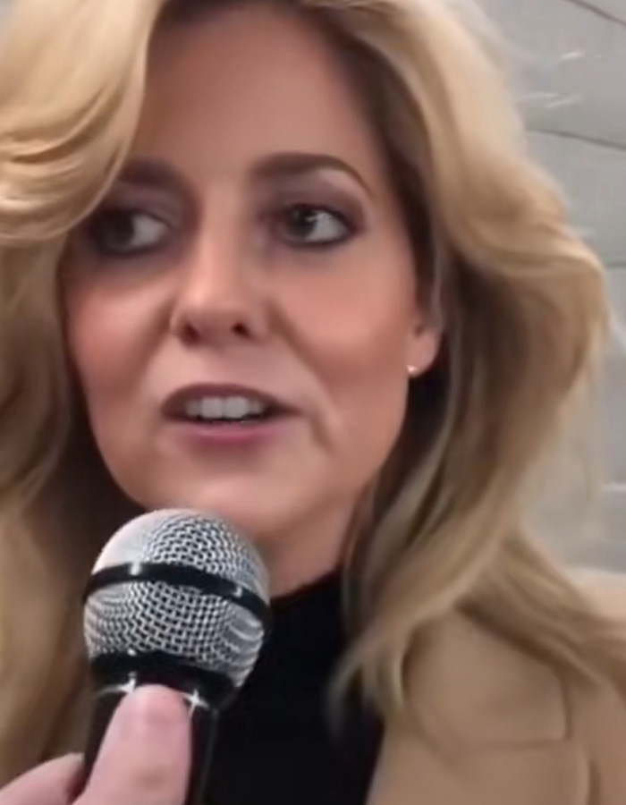 A blonde woman in a tan coat speaks into a microphone, sharing facts that went down 6 years ago.