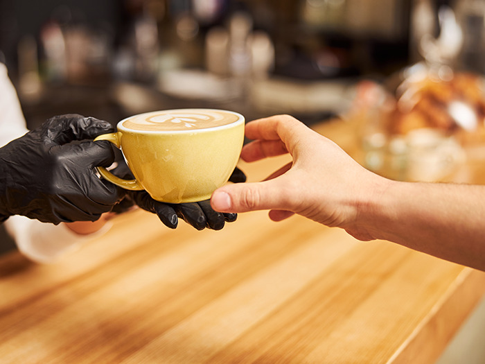A barista wearing black gloves handing a latte with foam art to a customer at a wooden counter showing rude customers learning.