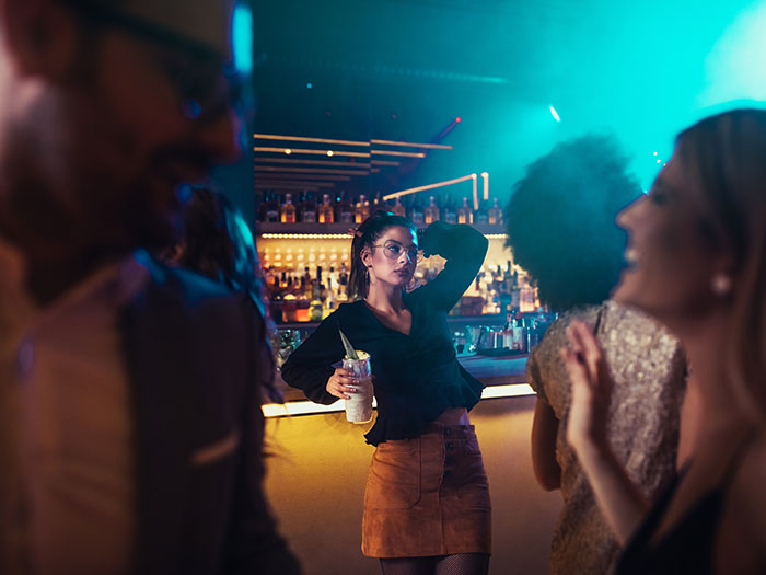 A woman in a club, looking away with a drink, possibly on a horrible first date. She is surrounded by other blurred people.