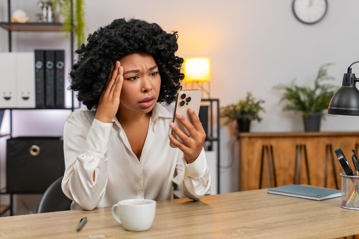 A woman with curly hair looks stressed, holding her phone and touching her forehead. Her expression mirrors the challenges of having celebrity relatives.