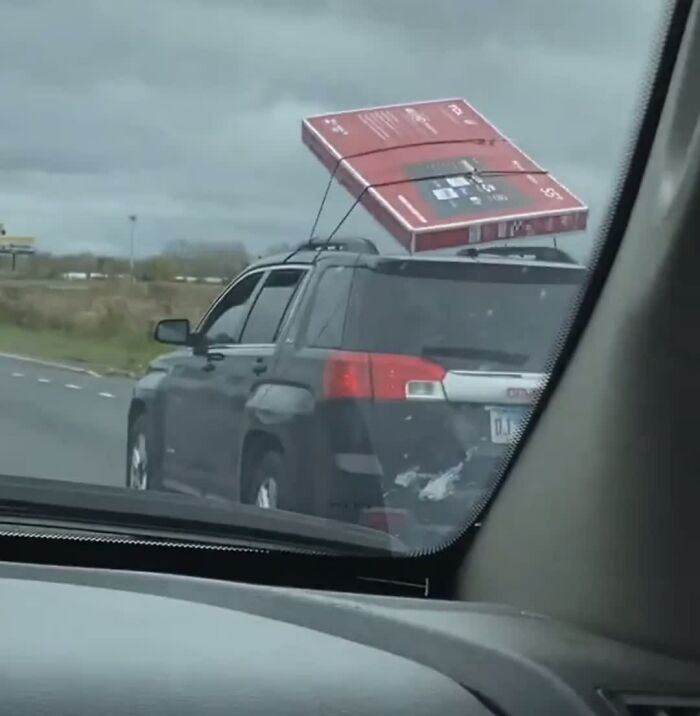 A car drives on the highway with a large, red boxed TV precariously strapped to its roof. A weird image of questionable transport.