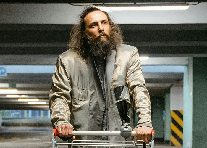 Man with unkempt hair and beard wearing a worn jacket, pushing an empty shopping cart in a dimly lit parking garage showing economy signs.