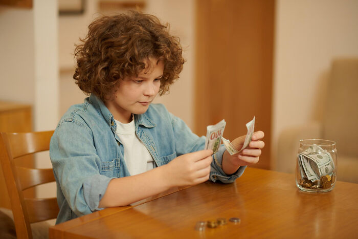 Curly-haired boy counting hundred-dollar bills, a jar of money on the table. A scene that could lead to friendship betrayals.