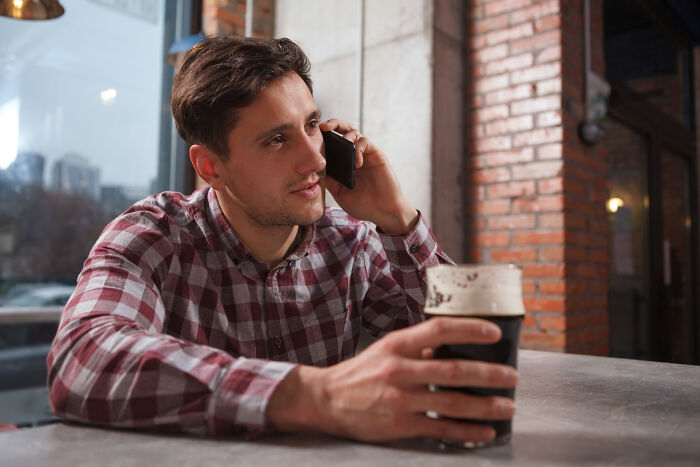 Young man in a plaid shirt holding a dark beer and talking on the phone, illustrating first dates with red flags and regret.