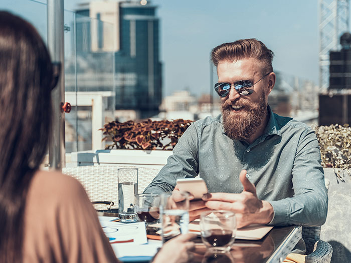 A man with a beard and sunglasses smiling, holding a phone across from a woman. A memorable first date moment.