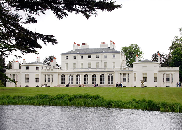 Large white mansion with people on the lawn near water, representing Harry and Meghan Markle turbulent dynamic location.