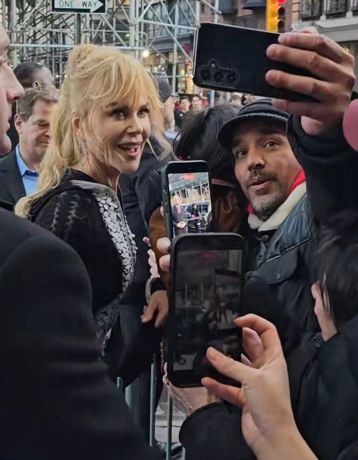 Nicole Kidman at a premiere, wearing a sheer outfit, smiling and posing for fans taking selfies on a busy city street.