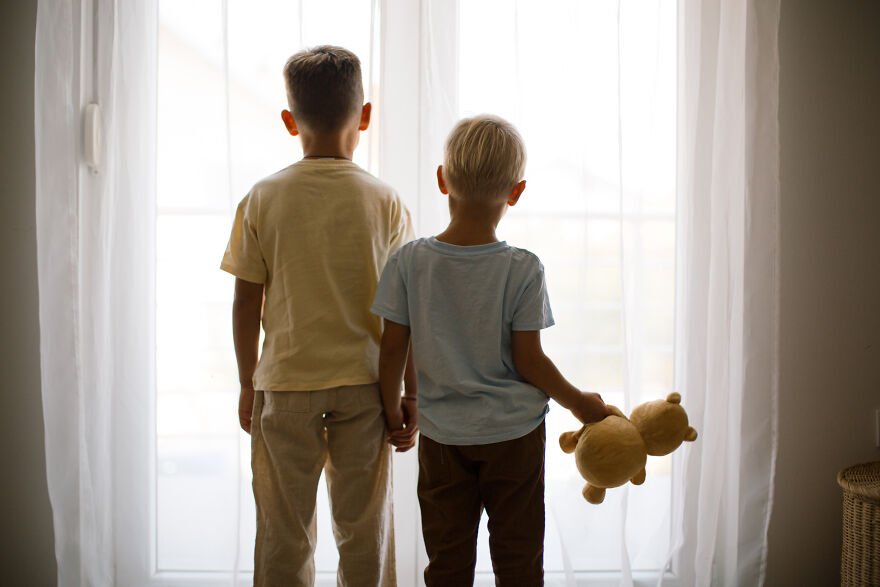 Two young boys holding hands, one with a teddy bear, standing by a window illustrating golden child syndrome concept.
